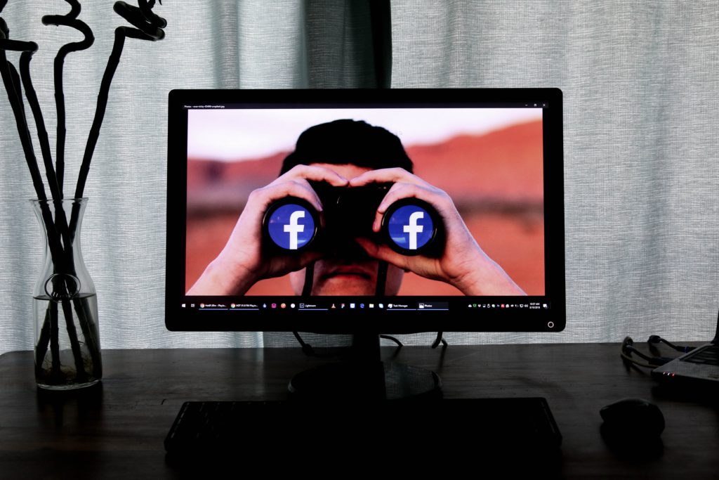 A high tech monitor, on a computer dark desk, in a calm and cosy dark room, displays a man holind binoculars and whatching facebook numbers increasing